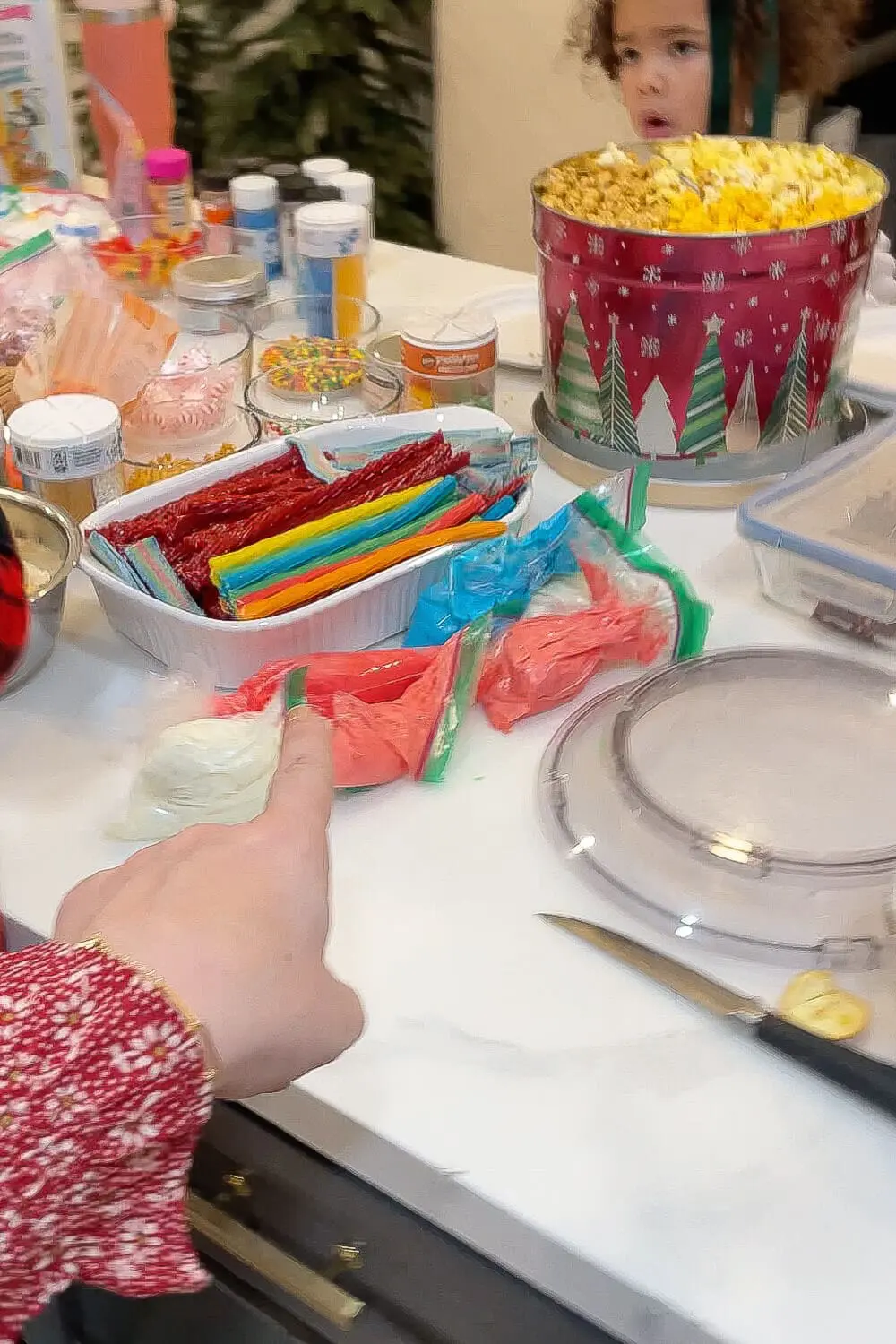 Close up of a gingerbread house candy station with piping bags of red, blue, green, and white royal icing, trays of licorice and rainbow candy strips, sprinkles, and a Christmas popcorn tin.