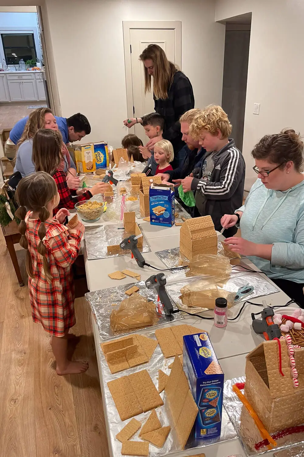 Family and friends gathered around a long table at a gingerbread house decorating party, with kids and adults building graham cracker houses, hot glue guns on foil-covered boards, and boxes of crackers and candy spread out.