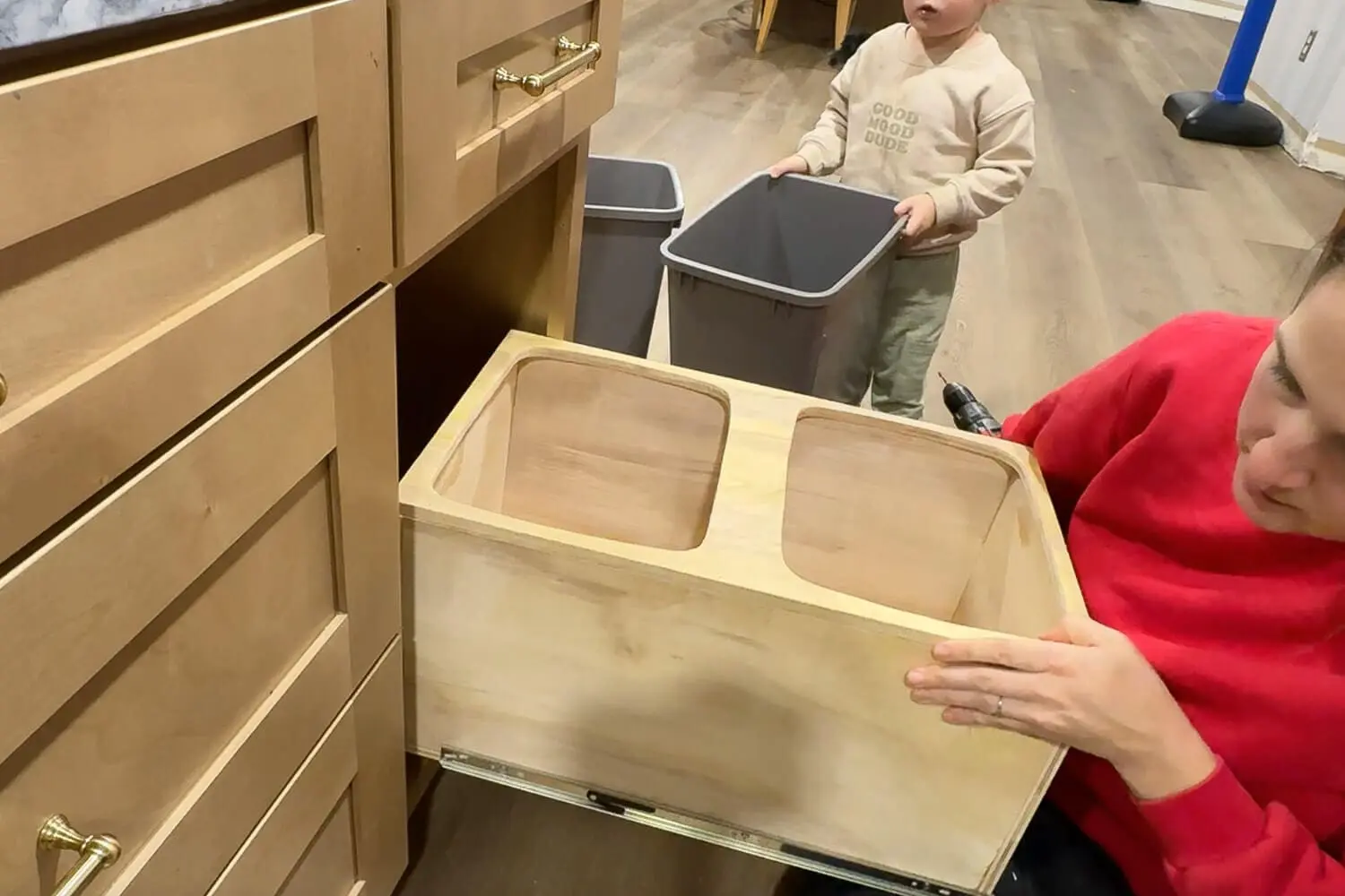 Sliding the finished plywood double trash can insert onto the drawer slides inside the base cabinet while a toddler holds one of the gray trash bins, installing a DIY pull out trash can cabinet in the kitchen.