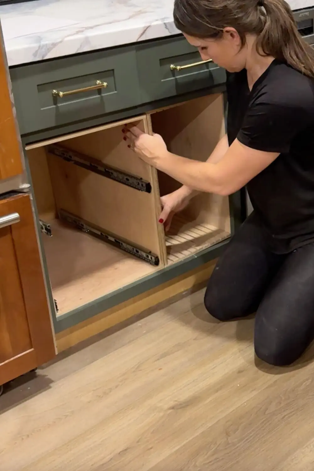 Woman sliding a newly built plywood pull-out drawer box with side-mounted slides into a lower kitchen cabinet to create a DIY organizer for food storage containers.
