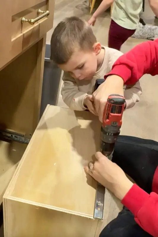 Little boy helping hold the drill while attaching drawer slides to a plywood box, building the pull out base for a DIY double trash can cabinet.
