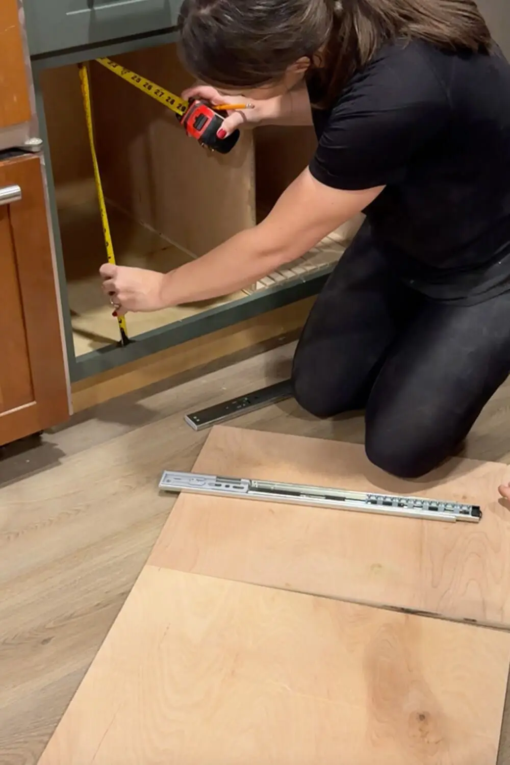 Woman measuring the inside of a lower kitchen cabinet with a tape measure while planning a custom plywood drawer organizer for glass food storage containers.