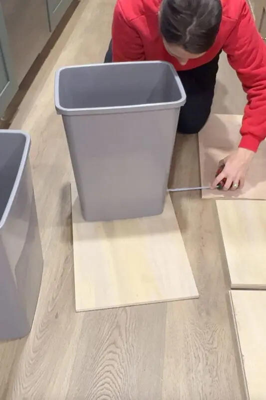 Measuring the spacing between two gray trash cans on a plywood base to size the platform for a DIY pull out double trash can cabinet.