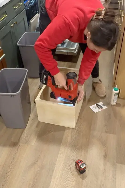 Using a cordless brad nailer to secure the top frame onto the plywood box, finishing the sturdy base for a DIY pull out trash can cabinet.