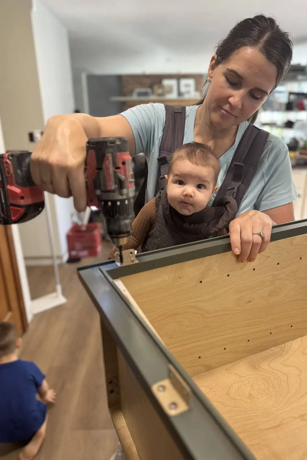 Assembling a new kitchen cabinet with a power drill while wearing a baby in a carrier, building the cabinet boxes during our DIY kitchen renovation.