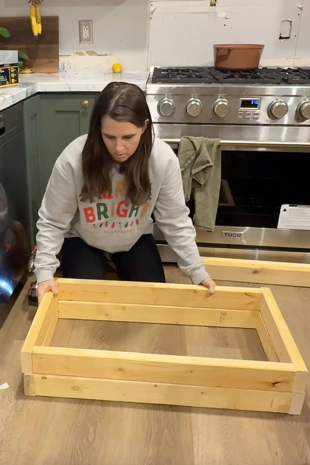 DIY builder assembling a rectangular wood frame on the kitchen floor for a DIY range hood cover, positioning cut boards and corner blocks before screwing the frame together in front of a stainless gas stove and dark green cabinets.