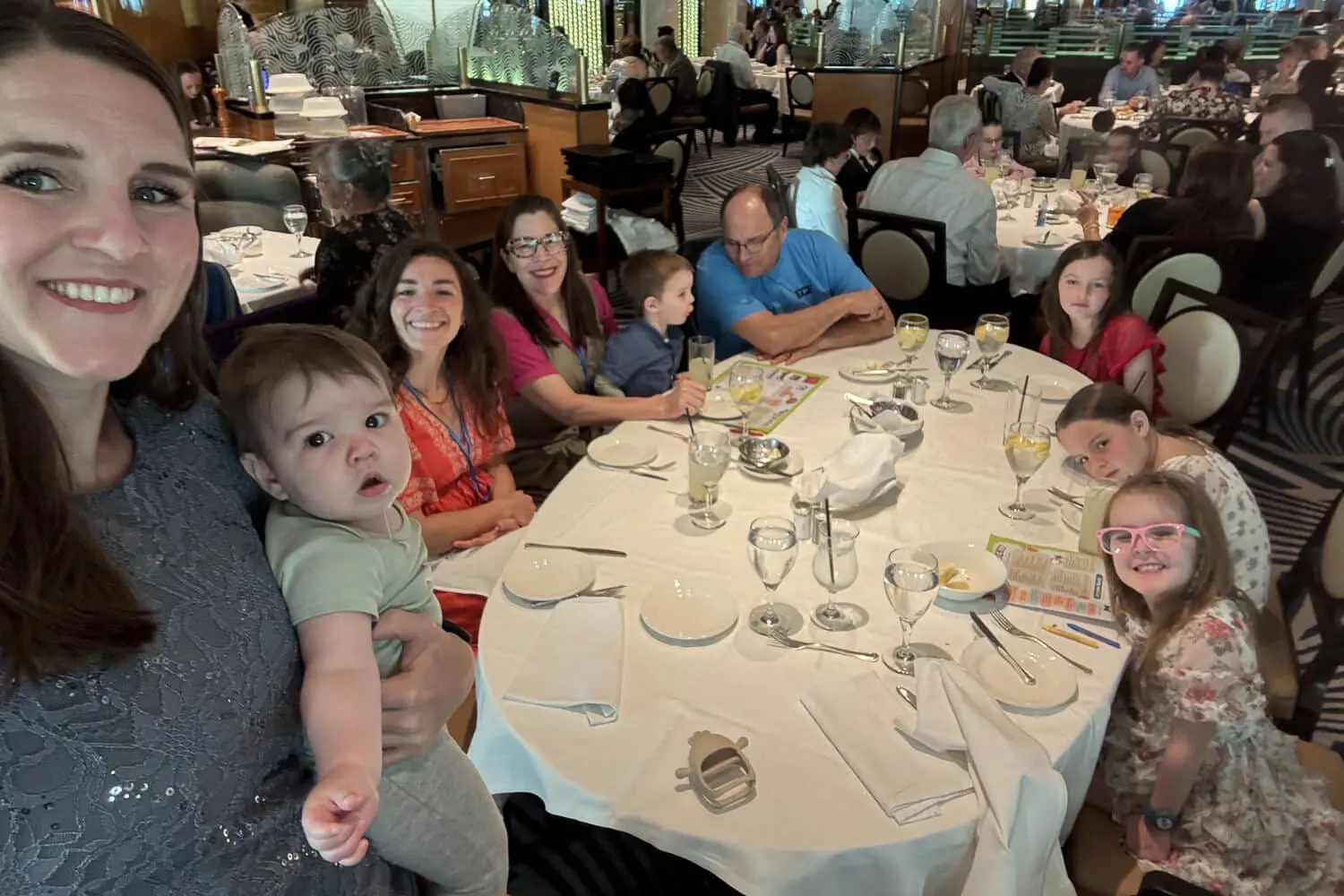 Big family sitting around a large round table in the cruise ship main dining room, with kids coloring on activity placemats and a mom holding a baby, showing what dinner looks like on a cruise with kids.