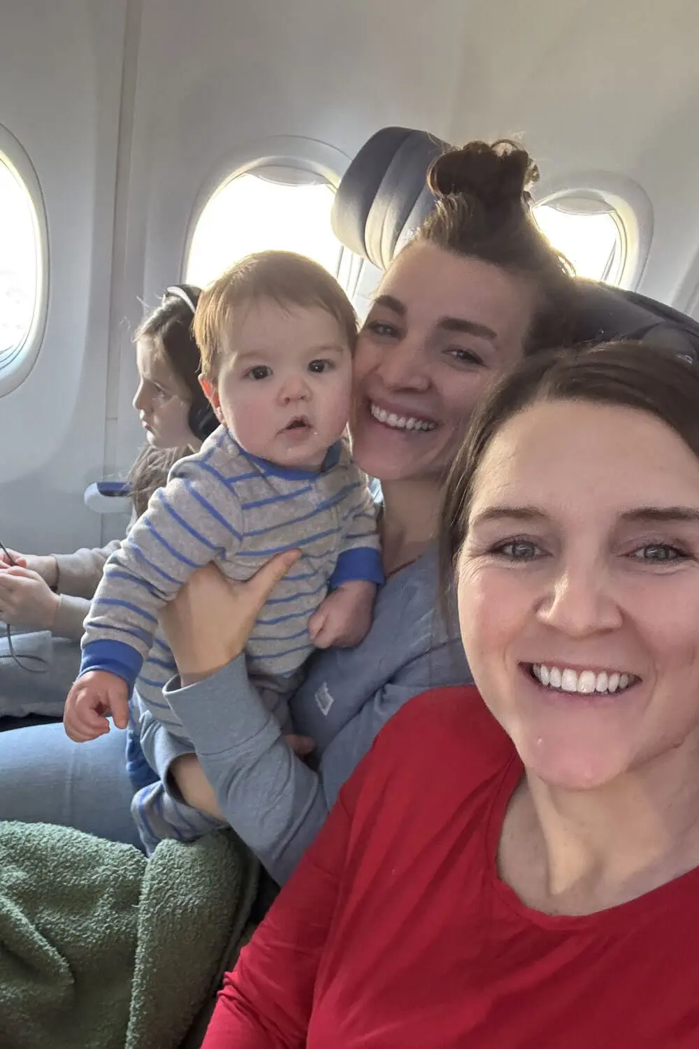 Airplane selfie on travel day for a vacation with young children, showing two women smiling in their seats while holding a baby in striped pajamas, with airplane windows in the background.