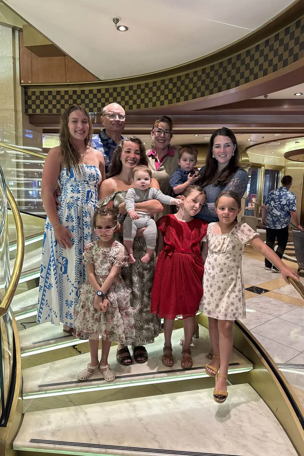 Multi-generation family posing on the lit atrium staircase aboard a Princess cruise, with gold railings, kids in dresses, a toddler, and a baby, on a cruise with kids trip.