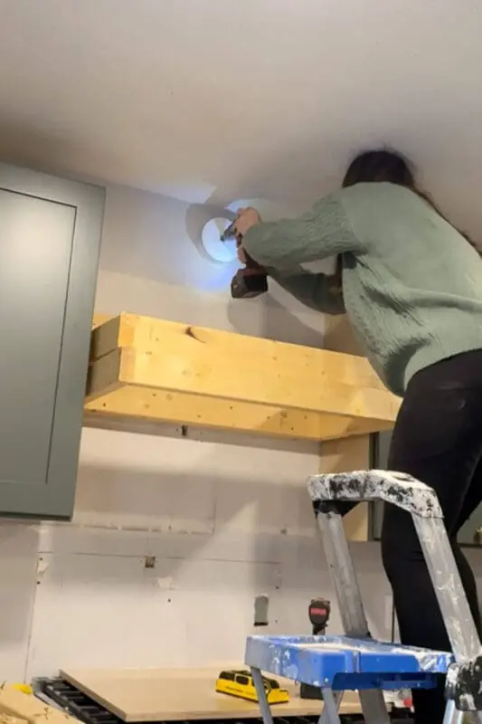 DIYer standing on a step ladder using a hole saw and drill to cut a circular opening for outside venting, routing the duct for exhaust.