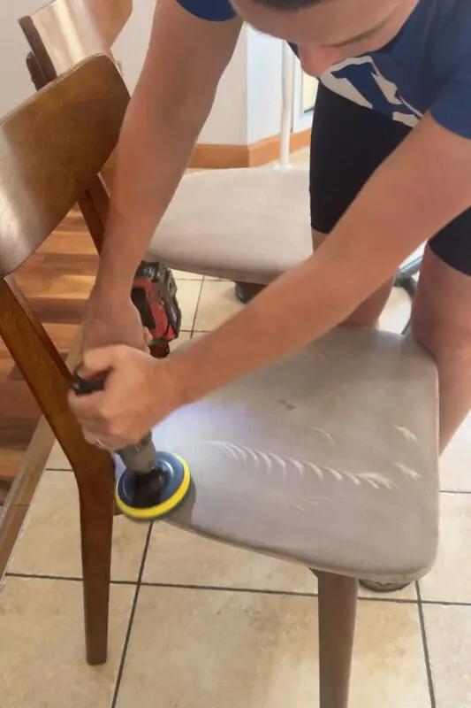 Using a drill brush to deep clean a stained upholstered dining chair seat, scrubbing out grime and set-in spots as part of a fabric chair cleaning tutorial.
