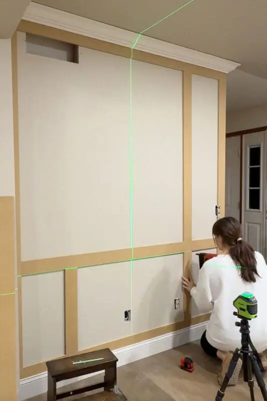 Building the DIY box molding wall trim grid by adding vertical trim pieces and smaller panels, using a laser level for even spacing.