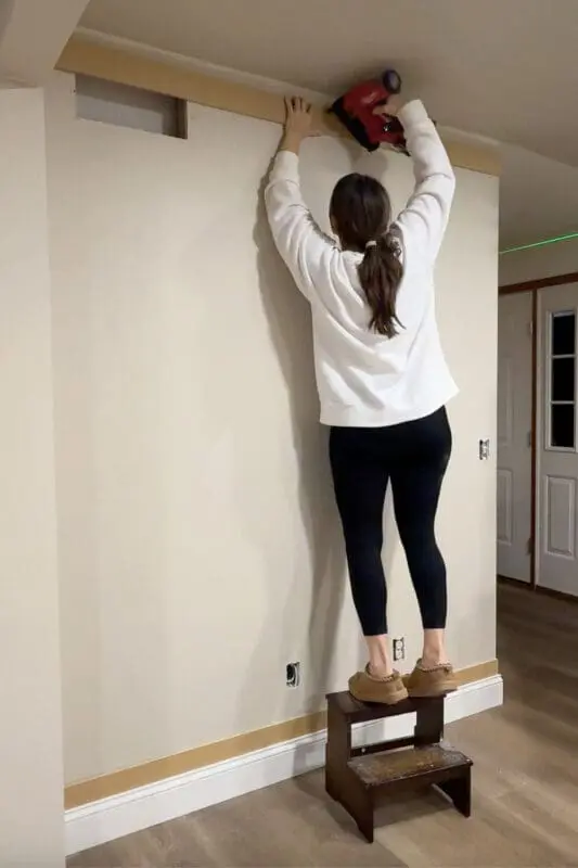 Installing the top trim board for DIY panel wall trim using a brad nailer, standing on a step stool for height.