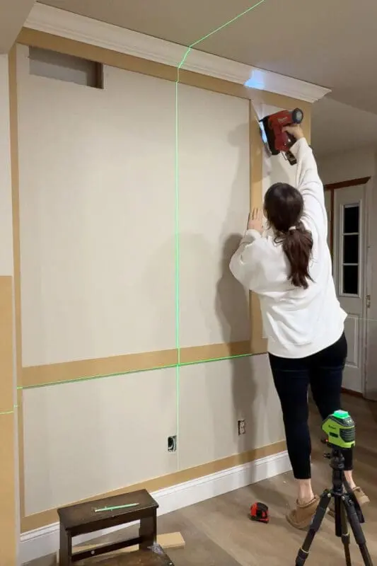 Installing a vertical divider for DIY box molding wall trim with a brad nailer while using a laser level to keep the layout perfectly straight.