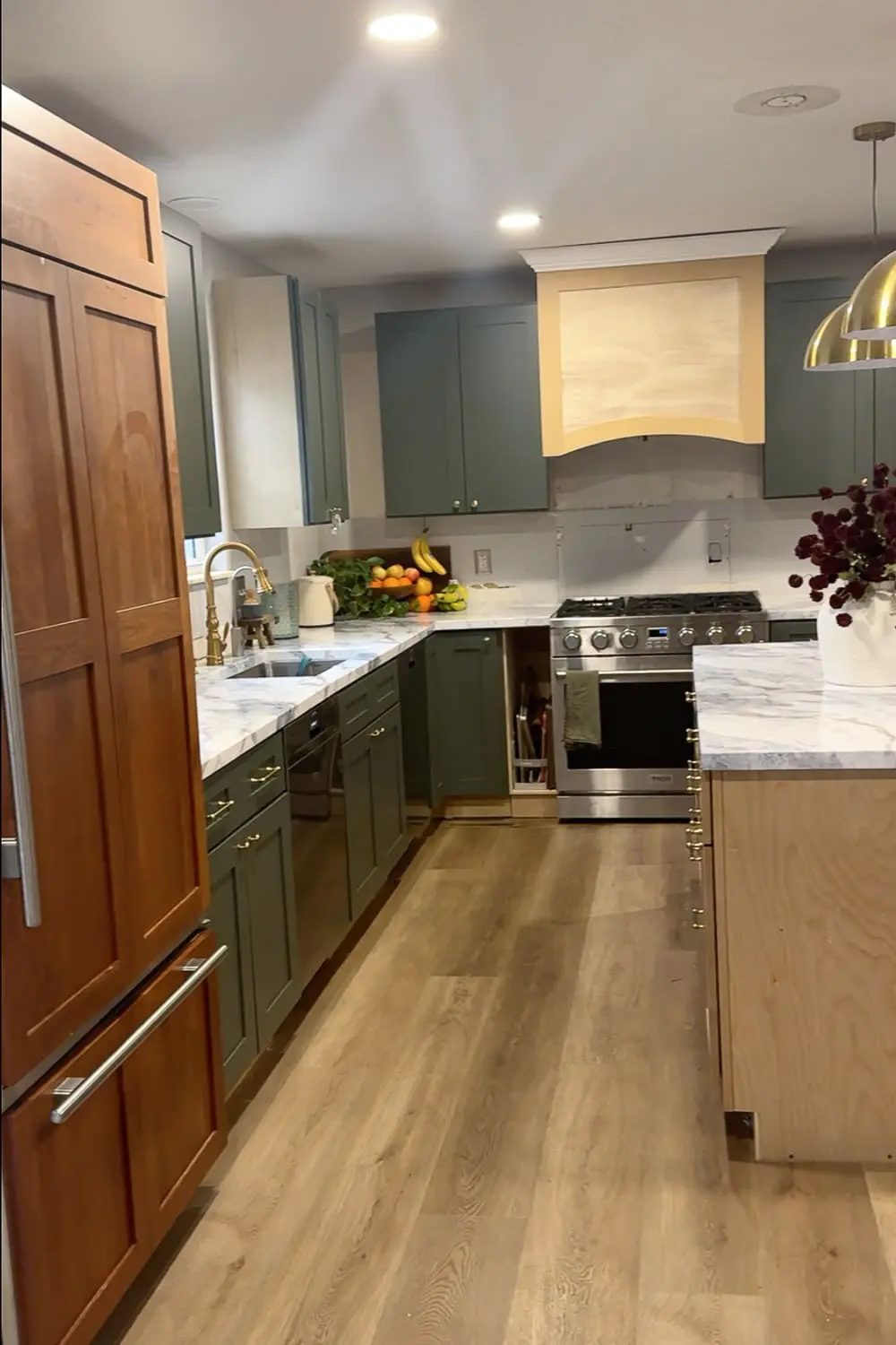Wide view of a DIY full kitchen remodel in progress with green cabinets, marble-look countertops, a wood island, brass hardware, and unfinished areas still being worked on.