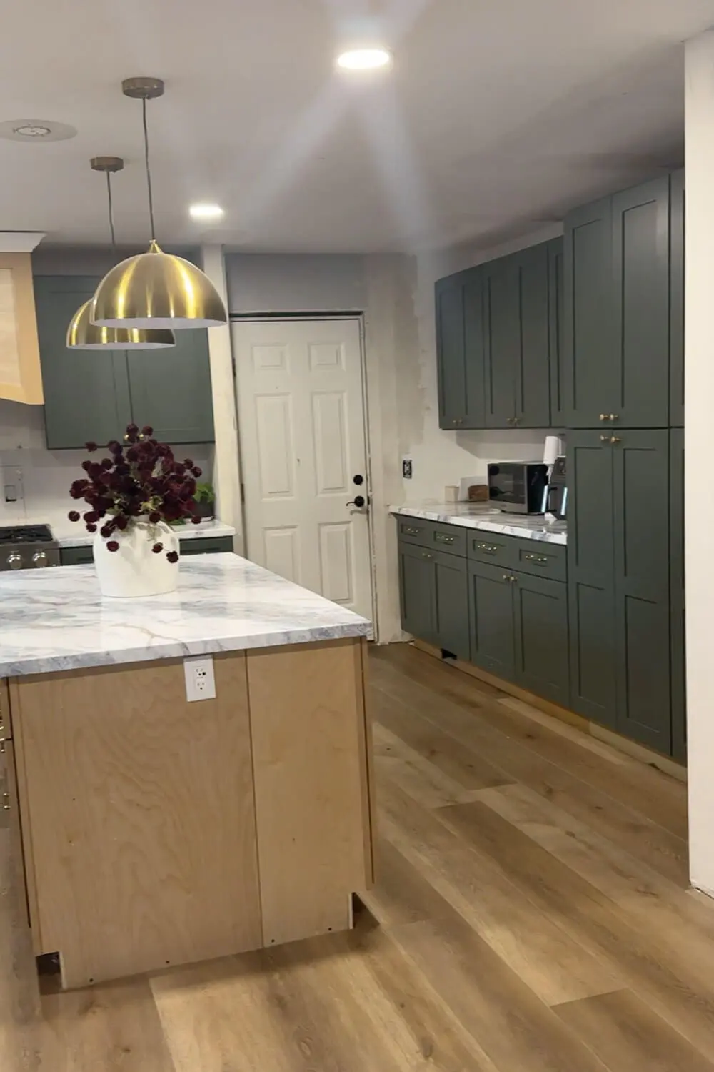 Current DIY kitchen remodel progress showing dark green cabinets, faux marble-look countertops, brass pendant lights, and an unfinished plywood island base.