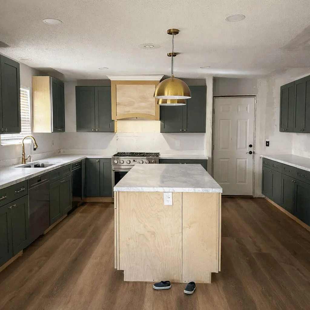 In-progress DIY kitchen remodel with dark green cabinets, a birch plywood island, white marble-look countertops cleared off, brass pendant lighting, and unfinished walls showing the remodel is still underway.