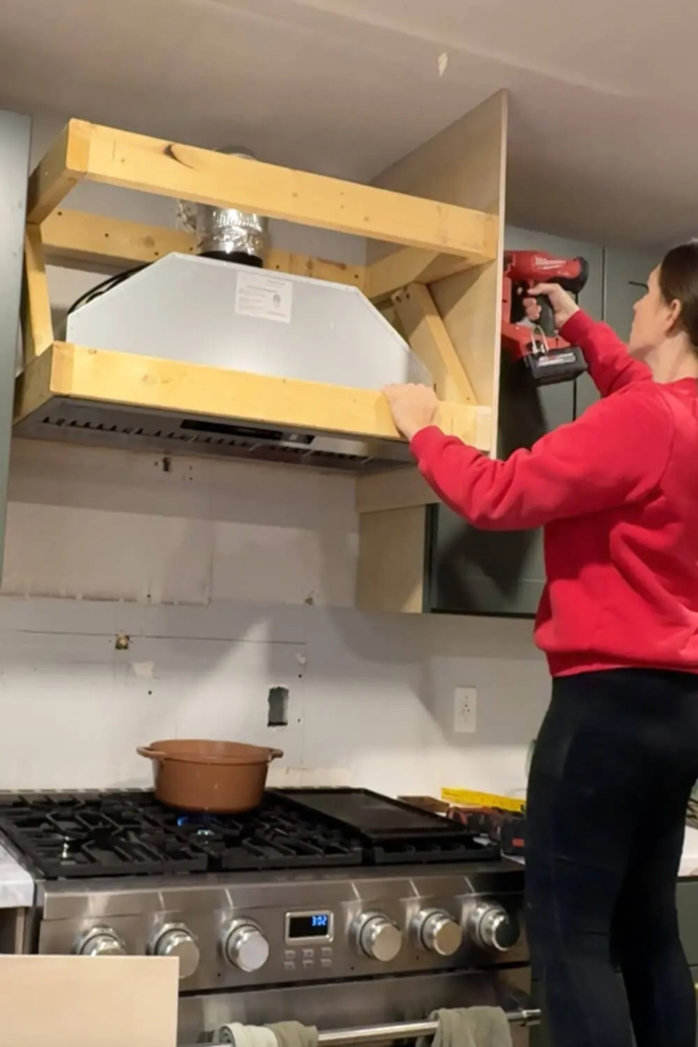 Attaching 1/4-inch plywood to wrap the DIY range hood cover frame over a stove, using a nail gun while the vent duct and hood insert are in place.