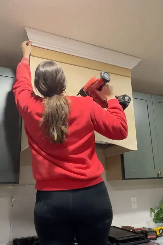 Installing crown molding on a custom plywood range hood cover, holding the crown in place and nailing it along the top edge for a built-in look.