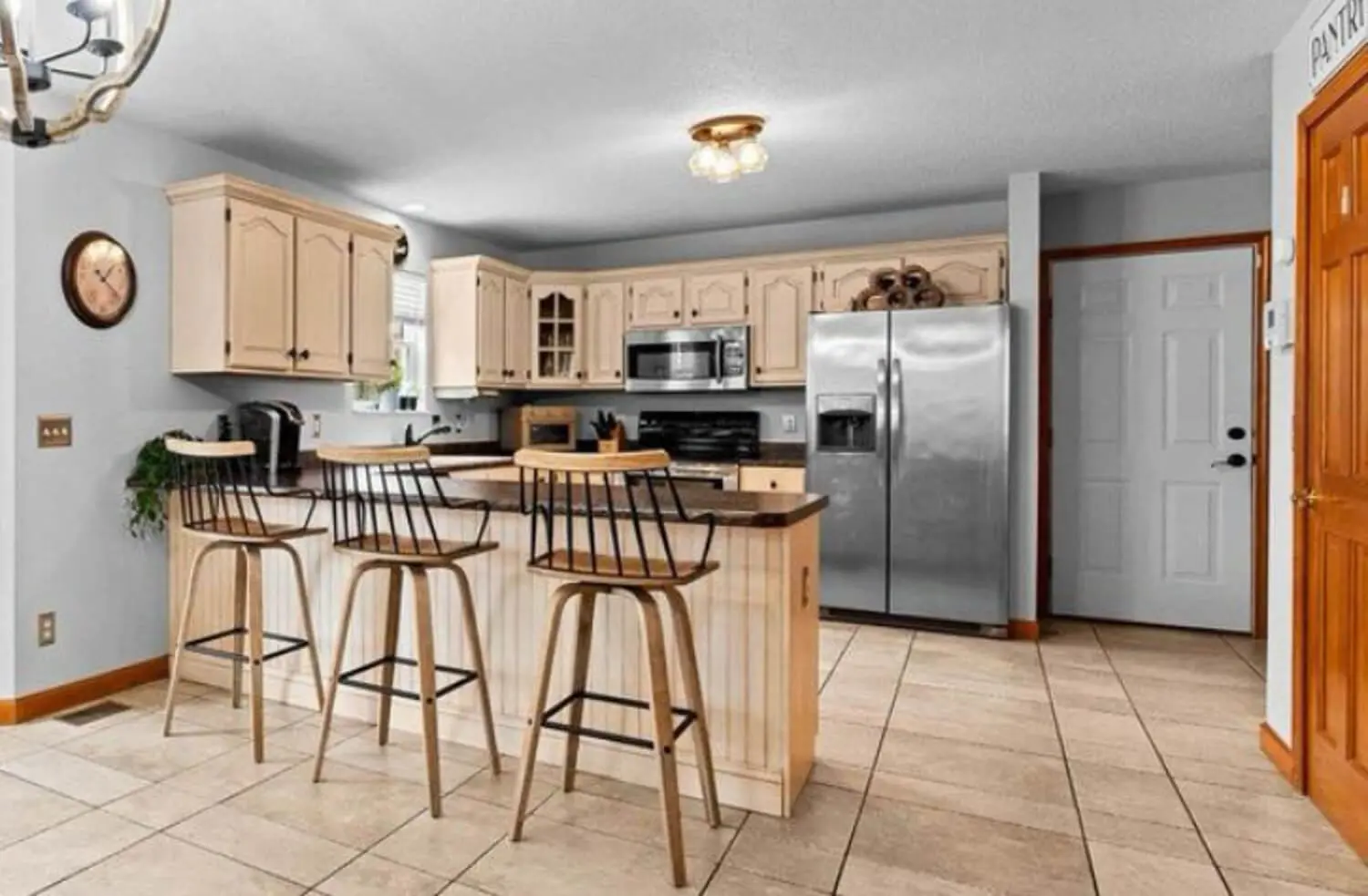 Dated room before renovation with beige raised-panel cabinets, stainless refrigerator and over-the-range microwave, white beadboard peninsula with three barstools, gray walls, and tan tile flooring.