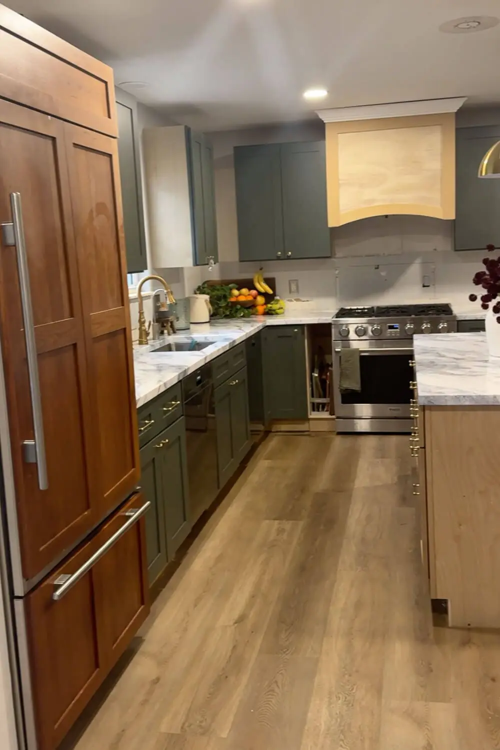 Current kitchen remodel view with dark green lower cabinets, faux marble-look counters, wood-look flooring, and the fridge panel area in progress near the island and range.