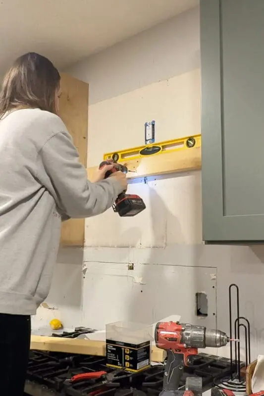 DIYer using a level while drilling 2x4 support boards into wall studs, adding solid blocking to securely attach the DIY wood range hood cover frame above the stove.