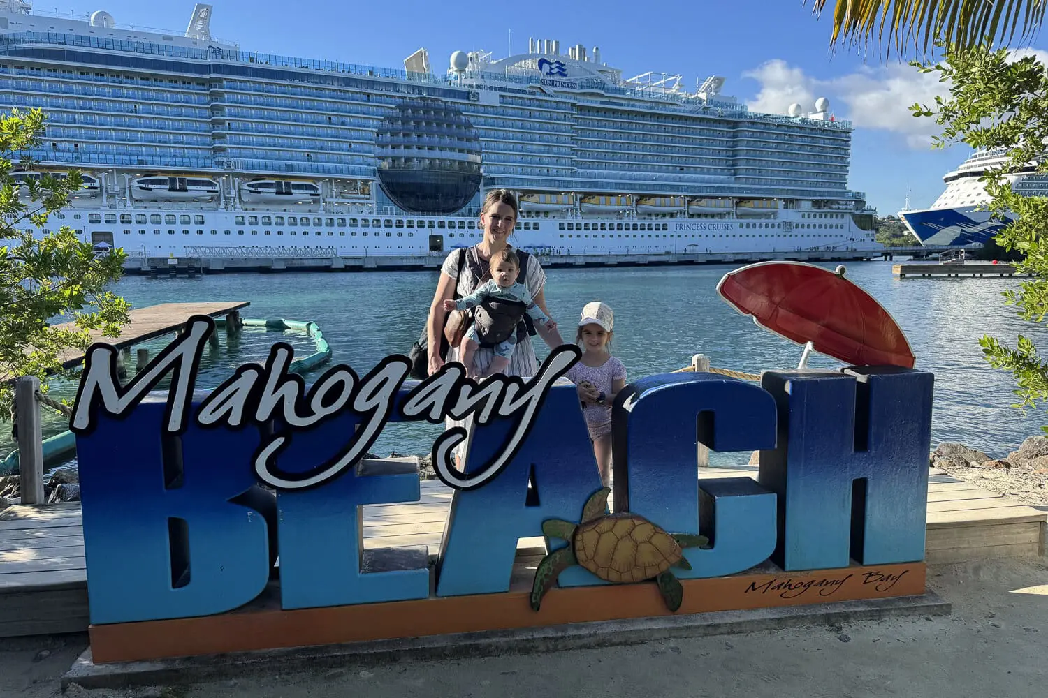 Mom wearing a baby in a carrier with a young child standing beside her at the Mahogany Beach sign in Mahogany Bay, Roatán, with a Princess Cruises ship docked in the background on a cruise with kids.