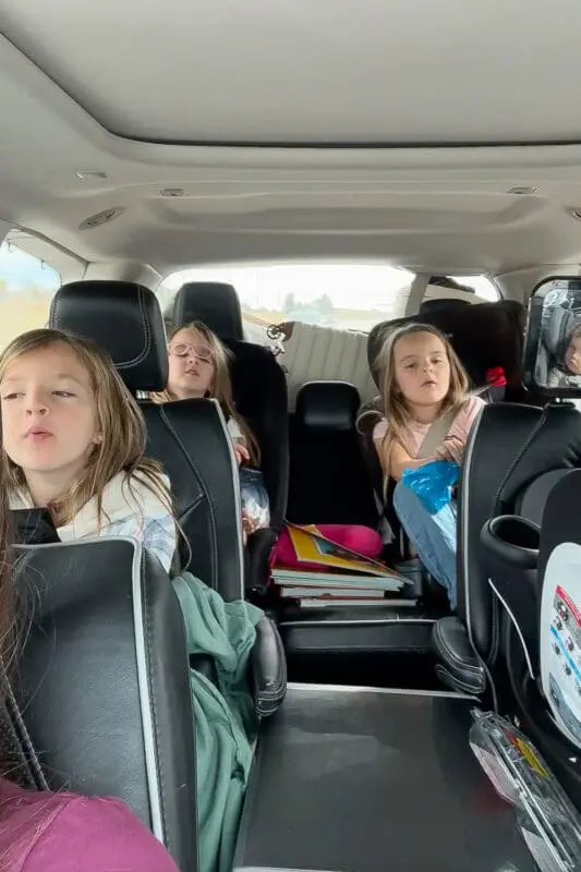 Kids strapped into car seats in the back of an SUV during a road trip with kids, with books and travel gear in the middle row, showing real-life road trip essentials for families.