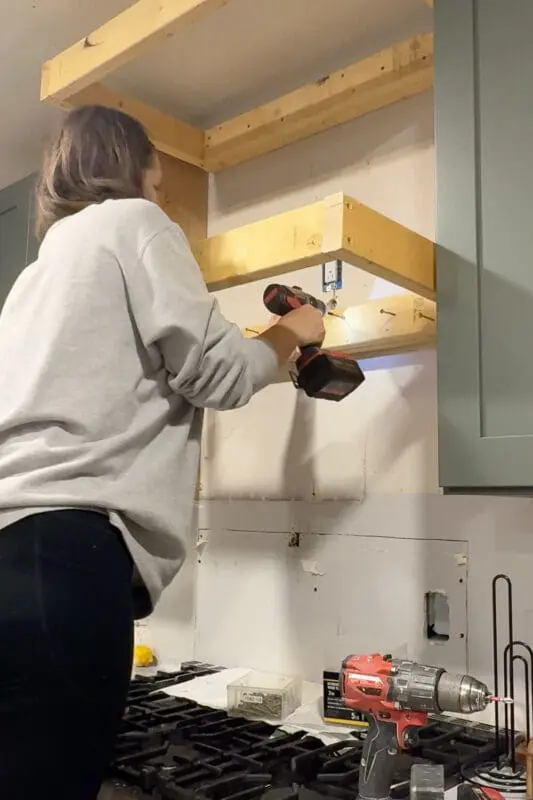 DIYer using a cordless drill to screw the wood range hood cover frame into 2x4 blocking and wall studs, attaching the sturdy base frame above the stove for a custom DIY wood range hood cover.