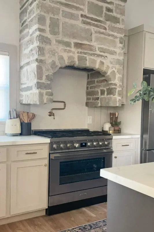 Stone-veneer range hood surround with an arched opening over a stainless range, paired with white cabinets, a brass pot filler, and simple neutral kitchen styling.