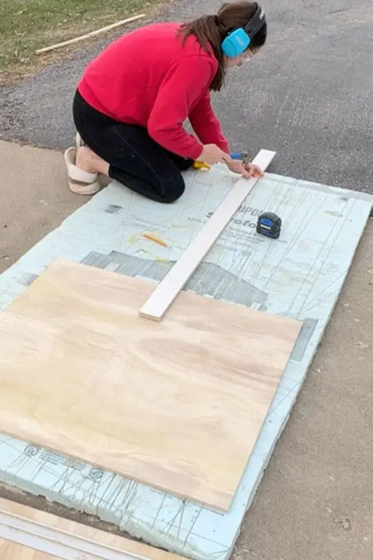 Kneeling on a foam board outside, using a piece of string and a straight board as a guide to mark an arched curve on a piece of plywood for a DIY kitchen project.