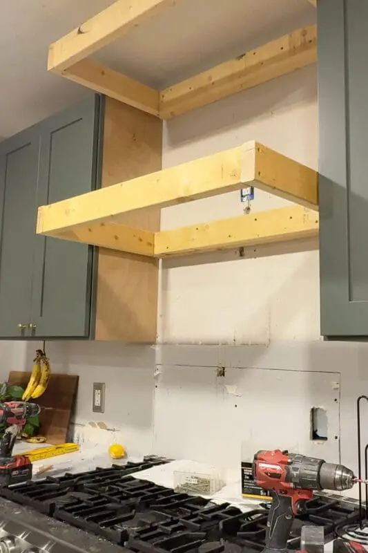 Wide view of the kitchen wall above the stove showing a rectangular wood frame test-fitted in place, aligned with 2x4 wall blocking to create the base structure for a DIY wood range hood cover between upper cabinets.