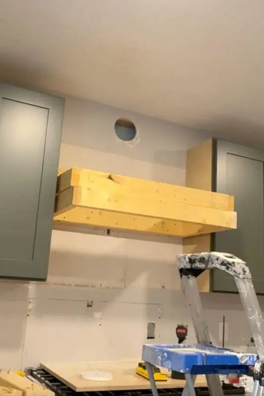 Kitchen wall above the stove showing a newly cut circular vent opening for a ducted range hood, positioned above the framed wood base for a DIY range hood cover during installation.