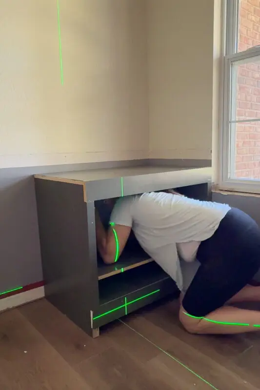 Attaching a finished DIY base cabinet to the wall using a laser level for alignment in a built-in cabinet project.