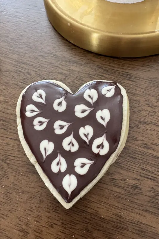 Heart-shaped sugar cookie flooded with dark royal icing without meringue powder and decorated with white piped heart designs on a wooden table.