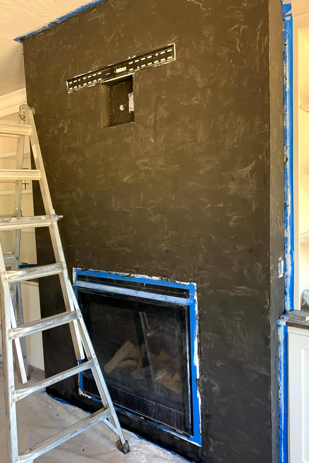 Fireplace wall fully covered in black textured plaster with TV mount bracket visible, blue painters tape still on edges, ladder nearby