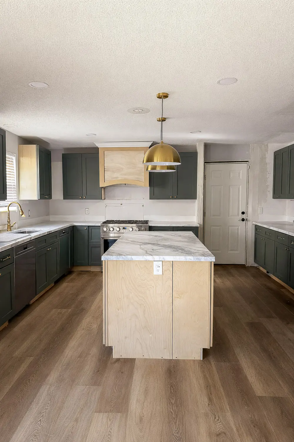 Wide view of a DIY kitchen remodel with a clean, clutter-free island featuring a faux marble epoxy countertop on a raw plywood base, green cabinets, brass pendant lights, and wood floors.