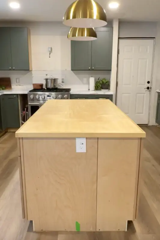 Front view of the DIY kitchen island with a raw plywood cabinet base and a plywood countertop installed, centered under brass pendant lights before finishing and painting.