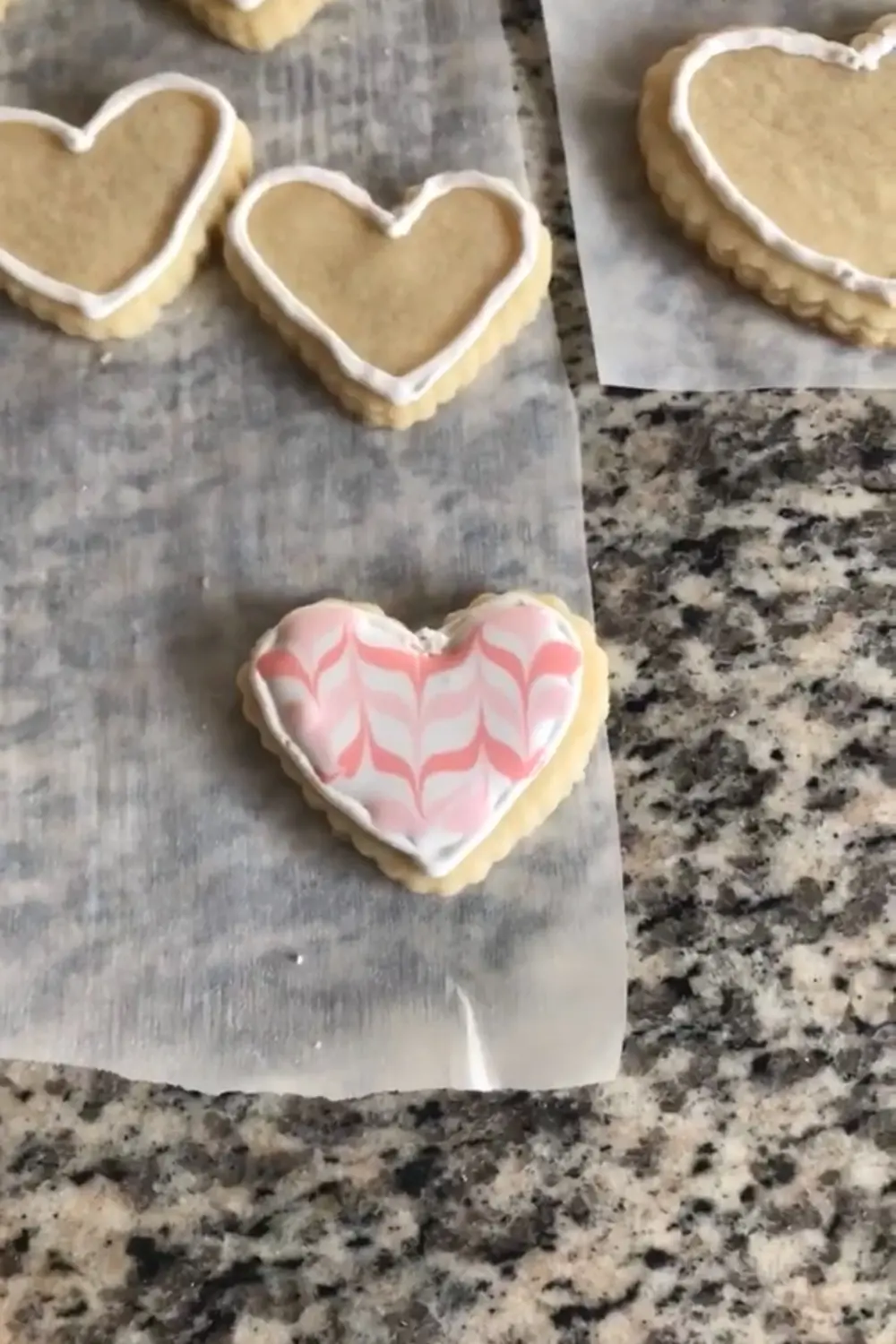 Heart-shaped sugar cookie decorated with a pink and white feathered royal icing pattern on parchment paper, with undecorated white-outlined heart cookies in the background.