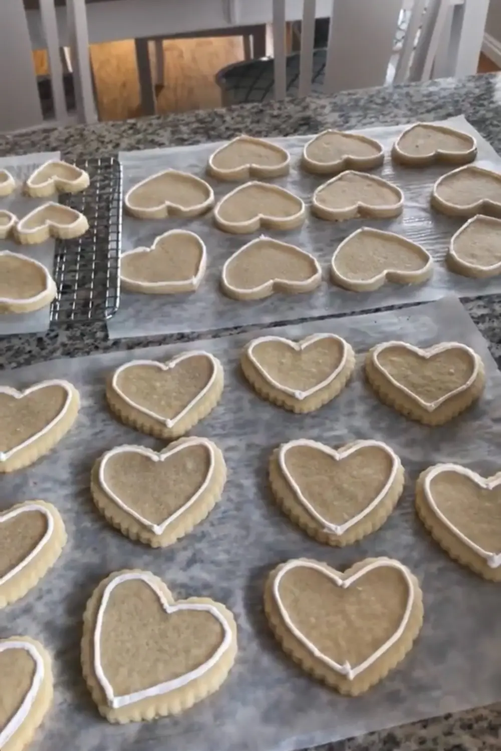 Heart-shaped sugar cookies laid out on parchment paper and a cooling rack, each outlined with white royal icing and ready to be flooded on a granite countertop.