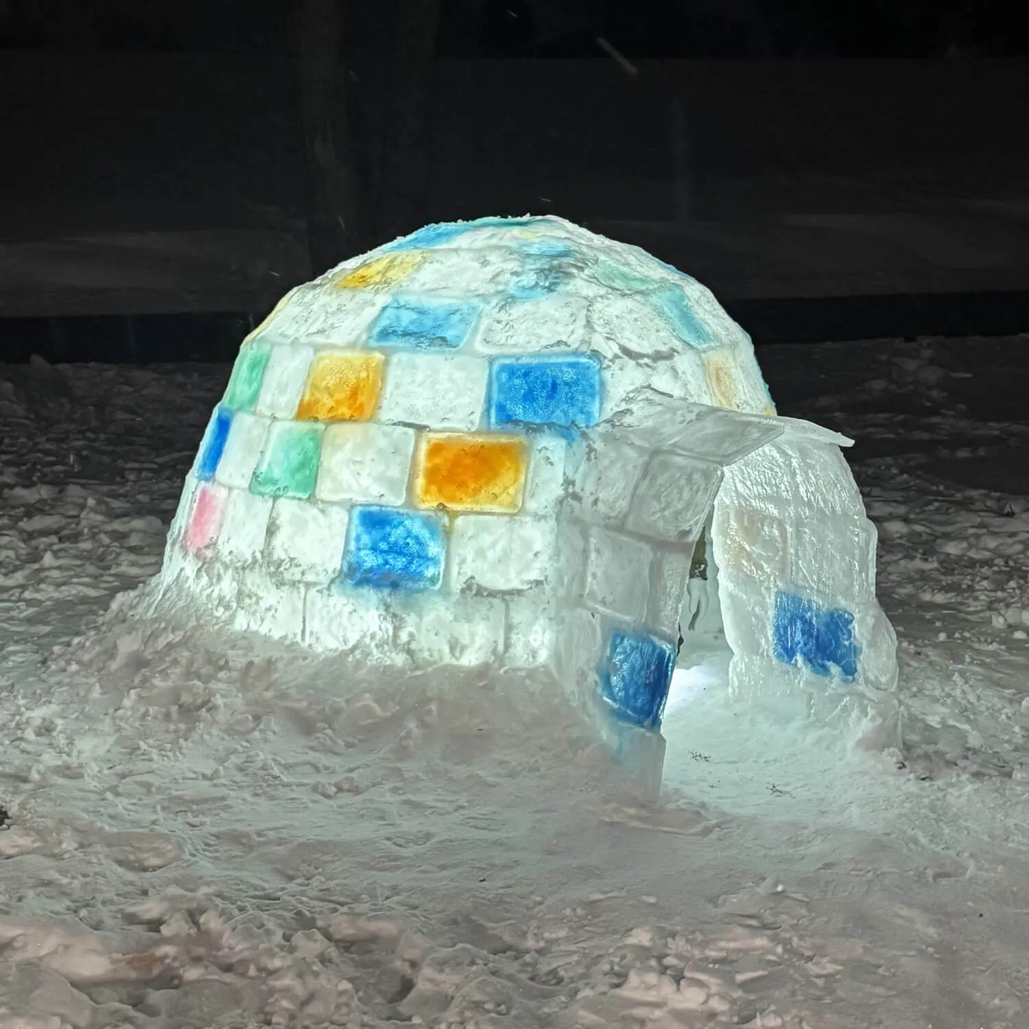 Close-up of a homemade backyard igloo built from ice blocks, featuring colorful frozen blocks and an illuminated doorway at night for how to build an igloo step-by-step