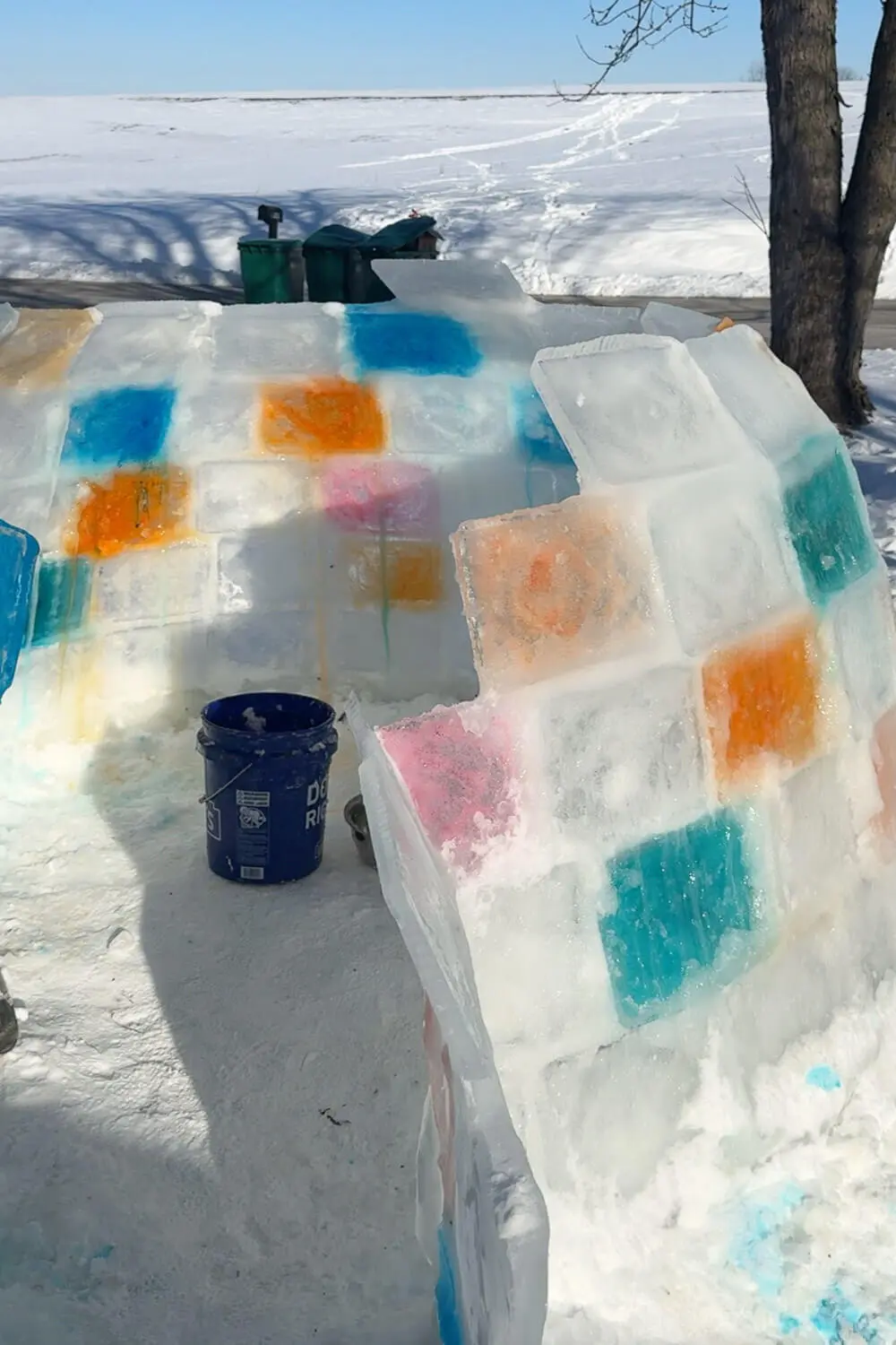 Daytime view of a backyard igloo in progress made from clear and colored ice blocks, showing the entrance opening and stacked walls before finishing the dome for a how to build an igloo tutorial