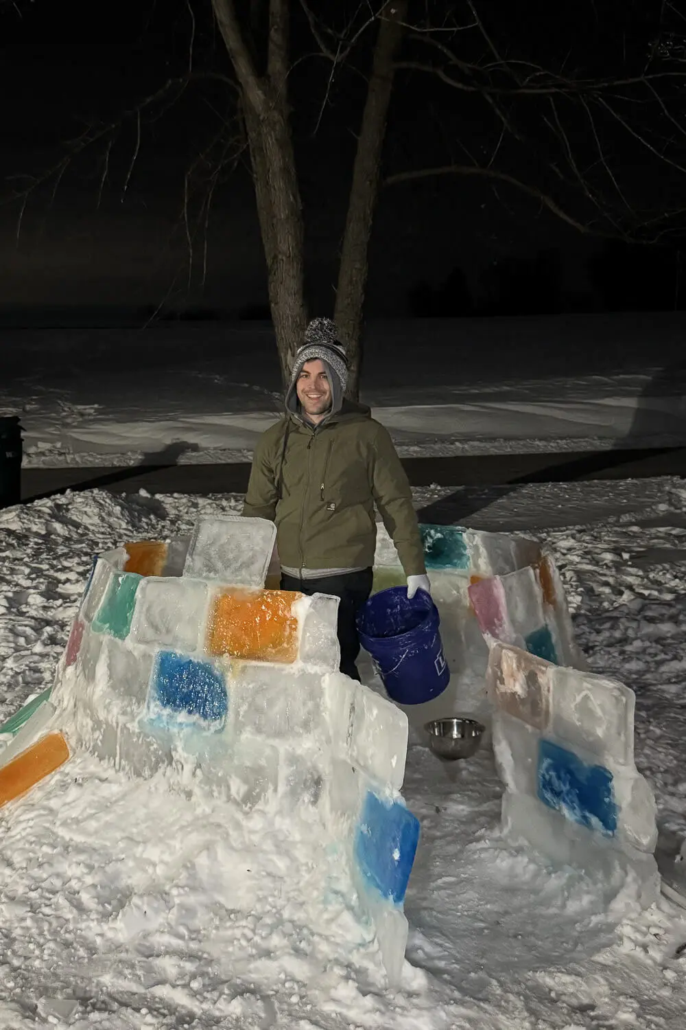 Backyard igloo in progress at night with stacked ice blocks and colorful frozen panels, showing the walls built up before the dome is finished in a how to build an igloo tutorial