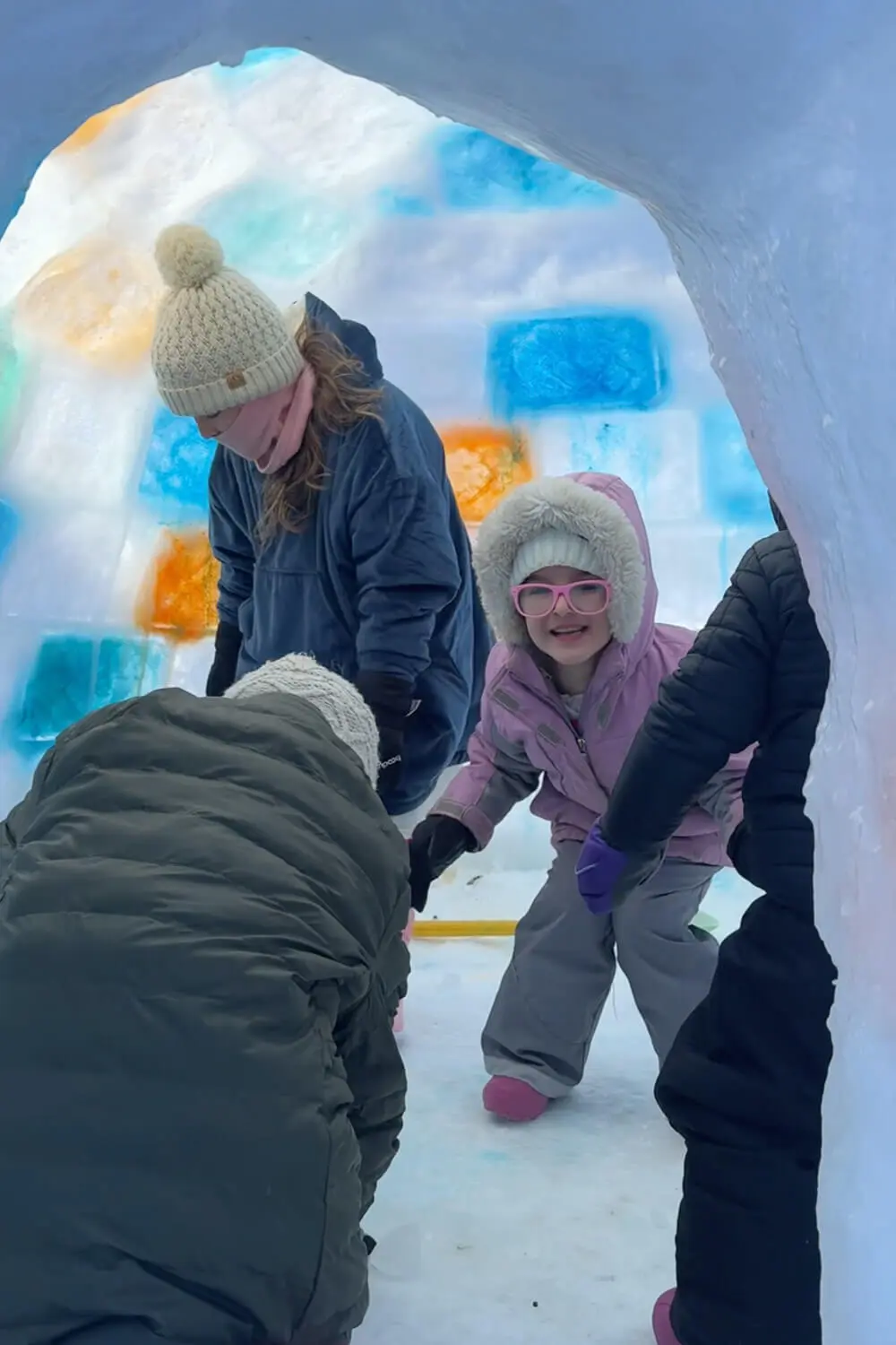 Kids and family inside a finished backyard snow fort made from clear and colored ice blocks, showing the arched entrance tunnel and glowing stained ice walls.