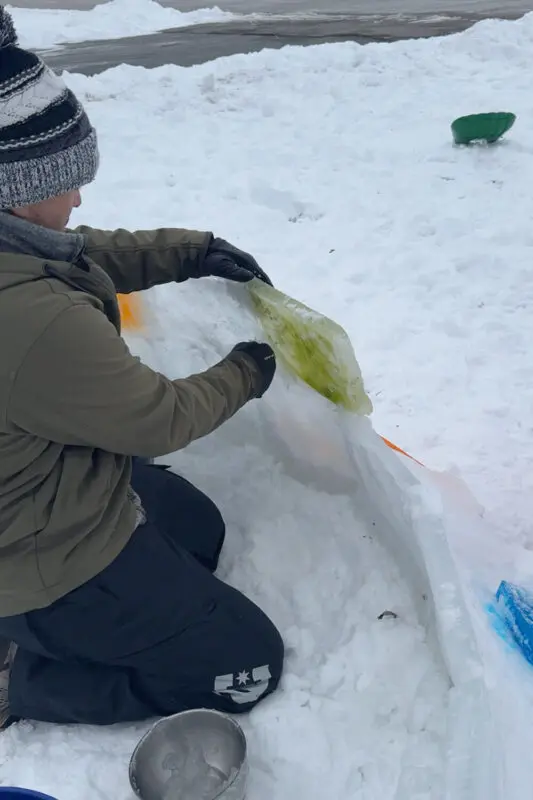 Adding the next colored ice block to the snow fort wall while packing snow-and-water slush into the seams to hold it together tightly.