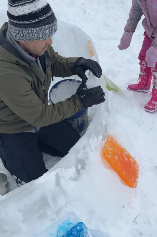 Spreading slush mortar on an igloo wall made from ice blocks, prepping the surface so the next block sticks, showing how to build an igloo step by step