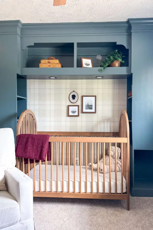 Nursery built-in crib wall painted deep blue with arched shelving, plaid wallpaper backing, framed art prints, and a natural wood crib styled with a cozy blanket.