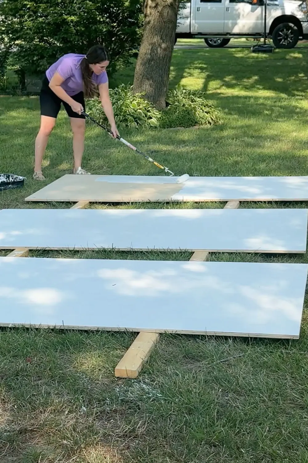 Rolling primer onto long plywood boards laid out in the yard, prepping the panels for painted DIY bookcases for our cozy home library.