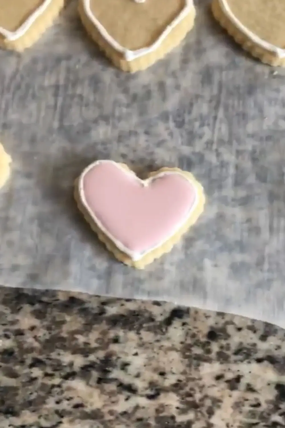 Heart-shaped sugar cookie fully flooded with smooth light pink royal icing on parchment paper, set on a granite countertop.