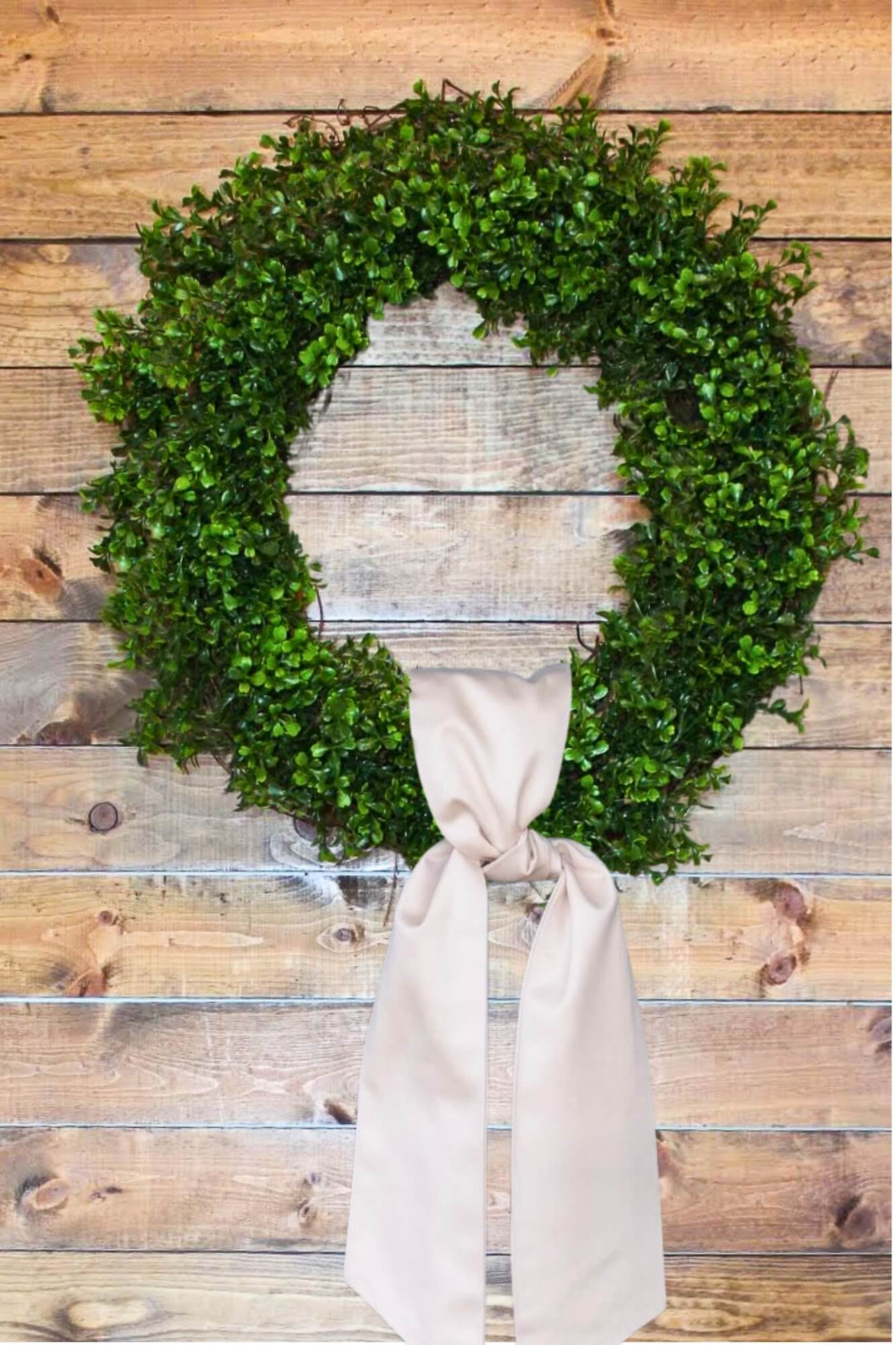 A finished DIY boxwood wreath with a large cream-colored bow hanging on a natural wood plank wall.